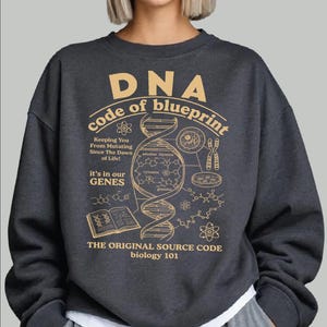 May include: A dark charcoal gray crewneck sweatshirt featuring a tan graphic print of a DNA double helix, chemical structures, and scientific text including "DNA code of blueprint," "it's in our GENES," and "THE ORIGINAL SOURCE CODE biology 101."