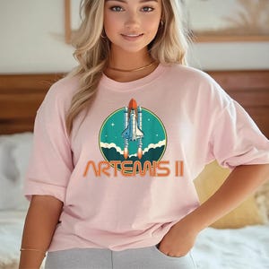 A woman wearing a pink t-shirt with a graphic of a space shuttle and the text "ARTEAIS II" printed on it.