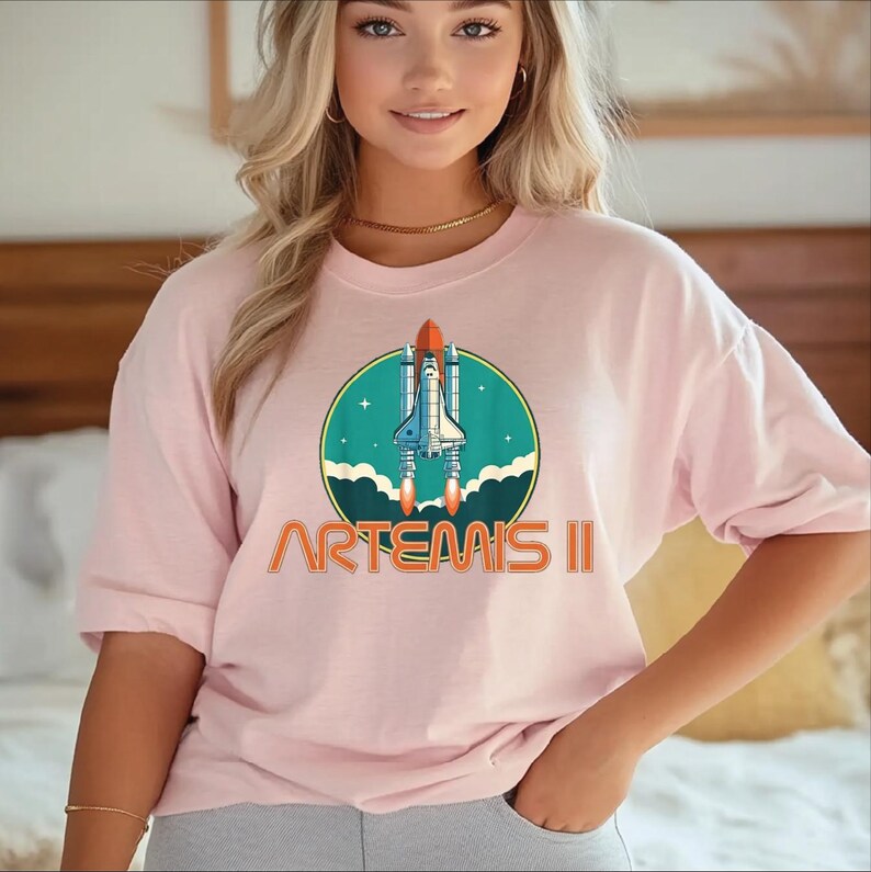 A woman wearing a pink t-shirt with a graphic of a space shuttle and the text "ARTEAIS II" printed on it.