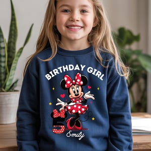 A young girl wearing a blue sweatshirt with a Minnie Mouse design and the text "Birthday Girl Emily" printed on it.