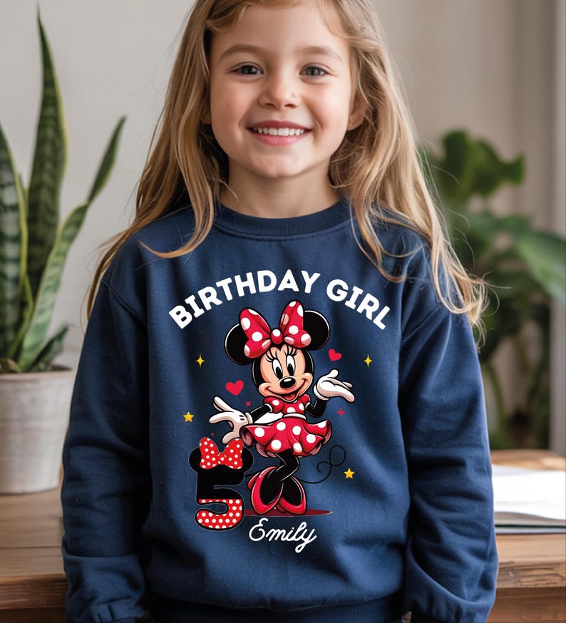 A young girl wearing a blue sweatshirt with a Minnie Mouse design and the text "Birthday Girl Emily" printed on it.