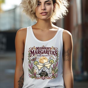 May include: A white tank top featuring a graphic print with the text "MIDNIGHT MARGARITAS" and "Moonlight, Magic & Sisterhood". The design includes two margaritas, a full moon, a moth, candles, and a house surrounded by floral elements.