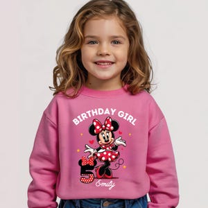 A young girl wearing a pink sweatshirt with a Minnie Mouse graphic and the text "Birthday Girl" printed on it.