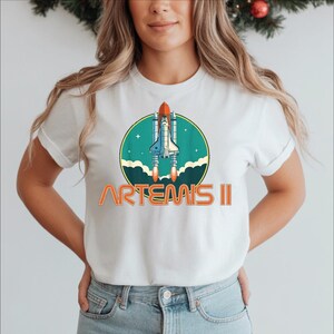 A woman wearing a white t-shirt with a graphic of a space shuttle and the text "ARTEMIS II" printed on it.