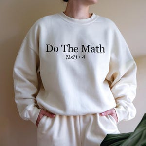 A person wearing a white sweatshirt with the text "Do The Math [9x7 + 4]" printed on it.