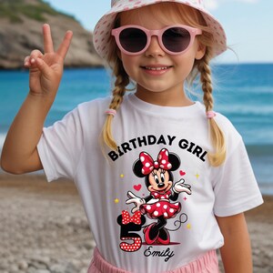 A young girl wearing a white t-shirt with a Minnie Mouse design and the text "Birthday Girl Emily" is smiling and making a peace sign.
