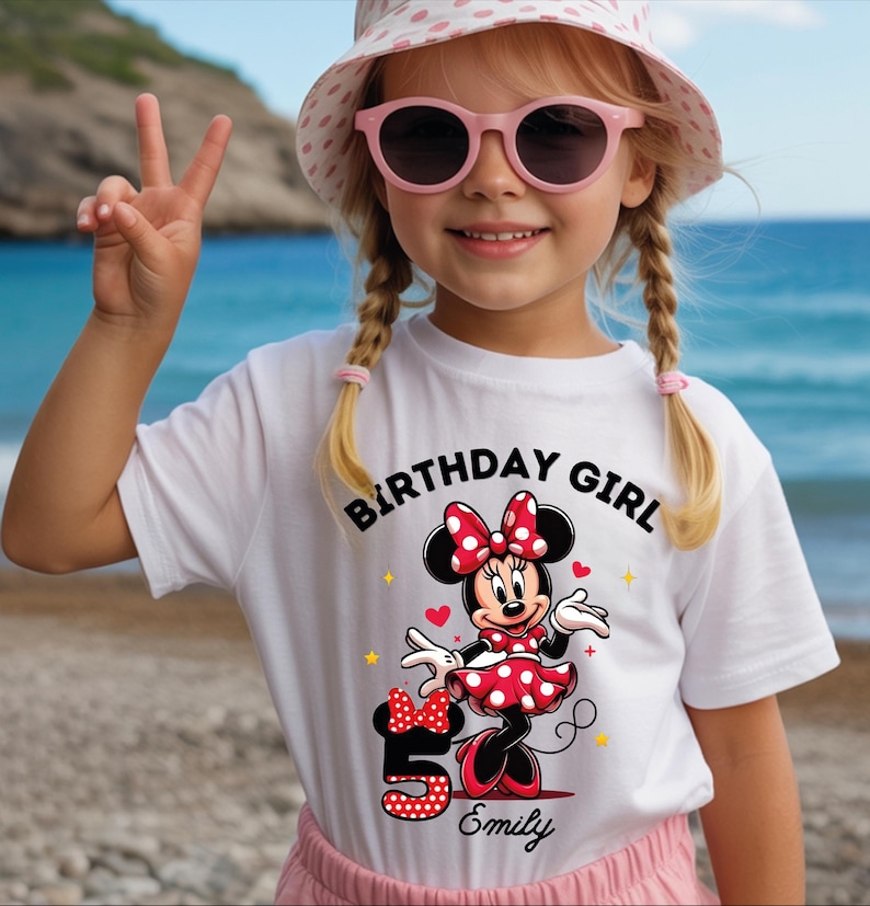 A young girl wearing a white t-shirt with a Minnie Mouse design and the text "Birthday Girl Emily" is smiling and making a peace sign.