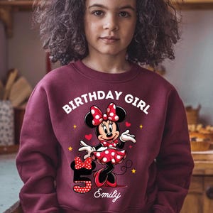 A young girl wearing a red sweater with a Minnie Mouse design and the text "Birthday Girl" printed on it.