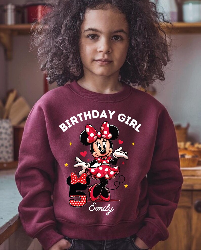 A young girl wearing a red sweater with a Minnie Mouse design and the text "Birthday Girl" printed on it.