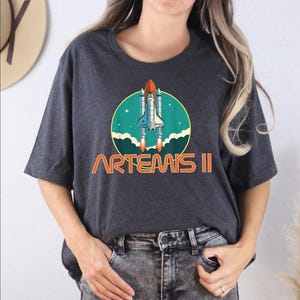 A woman wearing a gray t-shirt with a graphic design of a space shuttle and the text "ARTEMIS II" printed on it.