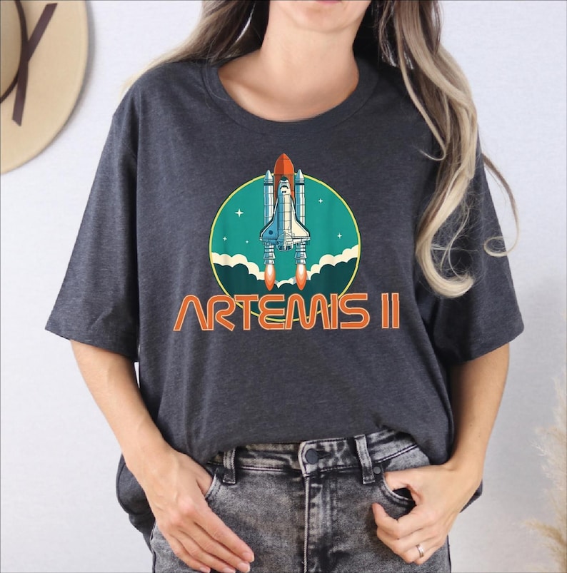 A woman wearing a gray t-shirt with a graphic design of a space shuttle and the text "ARTEMIS II" printed on it.