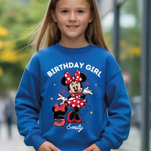 A young girl wearing a blue sweatshirt with a Minnie Mouse design and the text "Birthday Girl Emily" printed on it.