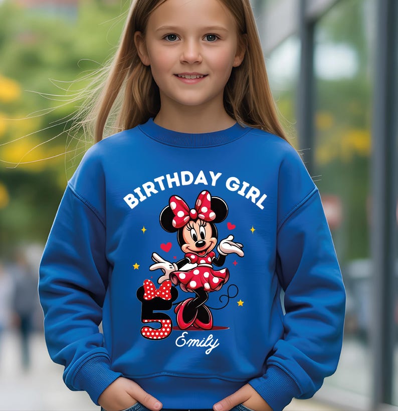 A young girl wearing a blue sweatshirt with a Minnie Mouse design and the text "Birthday Girl Emily" printed on it.