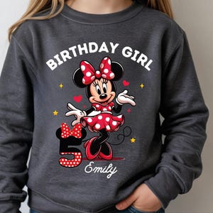 A young girl wearing a gray sweatshirt with a Minnie Mouse design and the text "Birthday Girl Emily" printed on it.