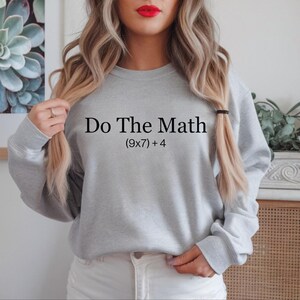 A woman with long blonde hair wearing a gray sweatshirt with the text "Do The Math [9x7] + 4" printed on it.