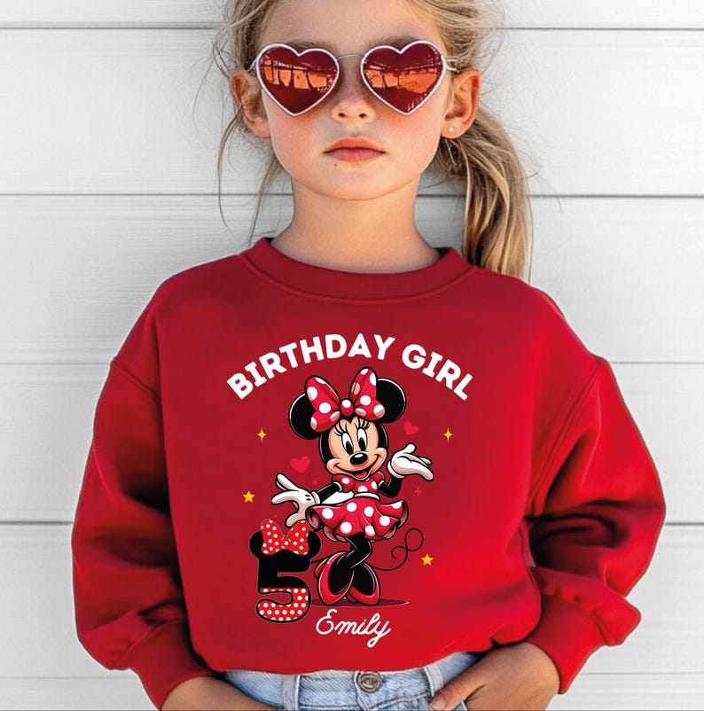 A young girl wearing a red sweater with a Minnie Mouse design and heart-shaped sunglasses, celebrating her 5th birthday.