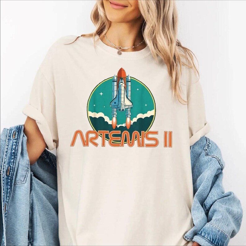 A woman wearing a white t-shirt with a graphic of a space shuttle and the text "ARTEMIS II" printed on it.