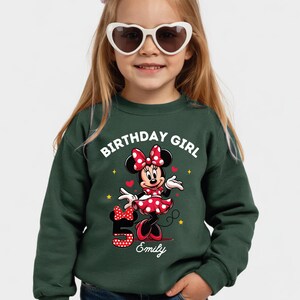 A young girl wearing a green sweatshirt with a Minnie Mouse design and the text "Birthday Girl" printed on it.