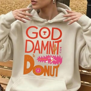May include: Cream-colored hoodie with a front graphic. The graphic features the words "GOD DAMNT DONUT" in orange and pink, along with various illustrations including a trophy, cactus, and a running figure. The hoodie has a front pocket and a drawstring hood.