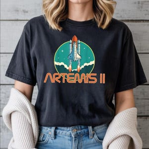 A woman wearing a black t-shirt with a graphic design of a space shuttle and the text "ARTEMIS II" printed on it.