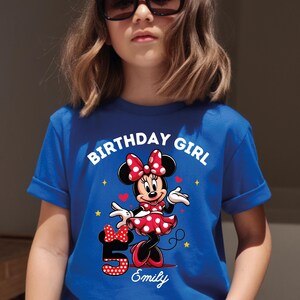 A young girl wearing a blue t-shirt with a Minnie Mouse design and the text "Birthday Girl Emily" printed on it.