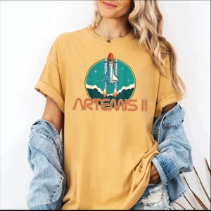 A woman wearing a yellow t-shirt with a graphic of a space shuttle and the text "ARTEMIS II" printed on it.