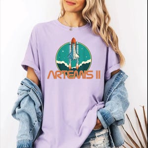 A woman wearing a purple t-shirt with a graphic of a space shuttle and the text "ARTEMIS II" printed on it.