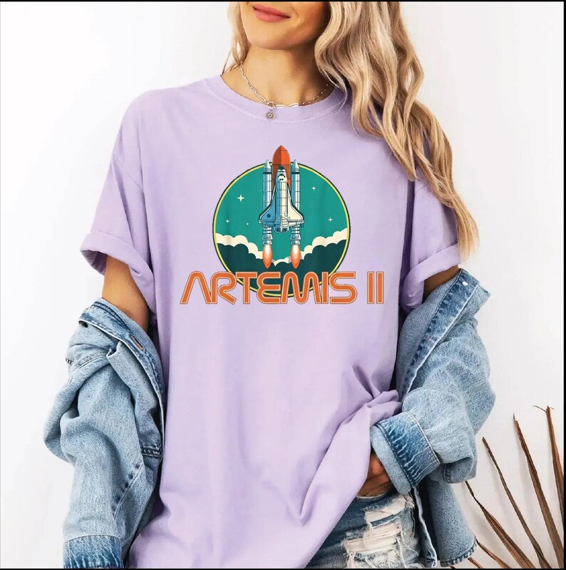 A woman wearing a purple t-shirt with a graphic of a space shuttle and the text "ARTEMIS II" printed on it.