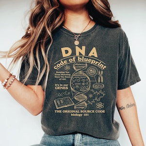 May include: A charcoal gray short-sleeve t-shirt featuring a tan graphic print of a DNA double helix, chemical structures, and an open book. The text reads DNA code of blueprint, Keeping You From Mutating Since The Dawn of Life, it's in our GENES.