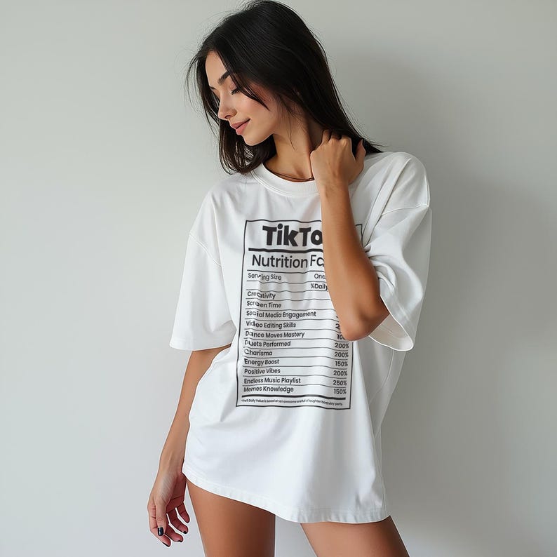 May include: White oversized t-shirt with a black graphic resembling a nutrition facts label. The label reads "Tik To Nutrition Facts" with various social media-related terms. The shirt is a casual, comfortable style.