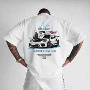 May include: White t-shirt featuring a graphic of two white sports cars with black accents. The text "Life is too short to drive boring cars" is above the cars. The shirt has a relaxed fit.