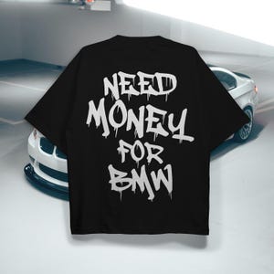 以下が含まれることがあります： 「NEED MONEY FOR BMW」と書かれた白いグラフィティスタイルのテキストが特徴の黒いTシャツ。Tシャツはベーシックなクルーネックです。白いBMW車が背景に一部写っています。