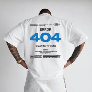 以下が含まれることがあります： 青色の「ERROR 404 HUMAN NOT FOUND」というテキストが入った白い半袖シャツ。シャツには、「UNICODE CODE ERROR」や「PREMIUM STREET WEAR」などの追加テキストも含まれています。