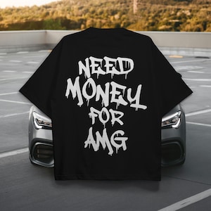 Pode incluir: Camiseta preta com texto branco "NEED MONEY FOR AMG" em estilo grafite. A camiseta é exibida ao ar livre, com um carro ao fundo. O texto tem um efeito de gotejamento.