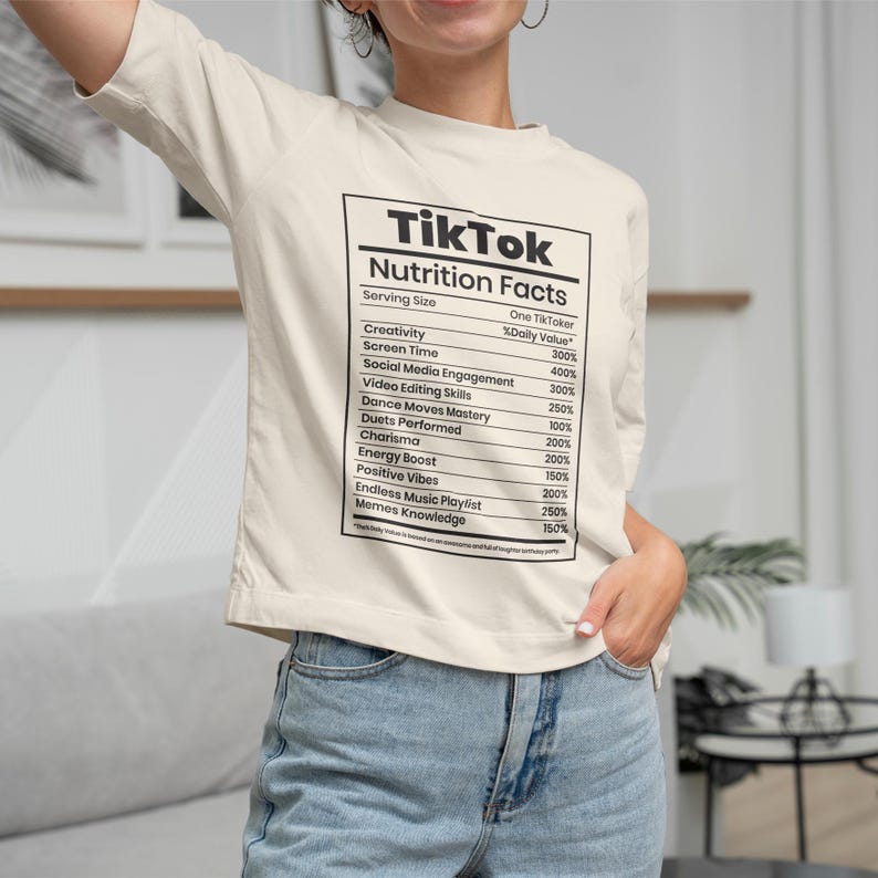 May include: Cream-colored t-shirt with a black and white graphic resembling a nutrition facts label. The label lists "TikTok" at the top, followed by various social media-related metrics and percentages. The shirt is worn with blue jeans.