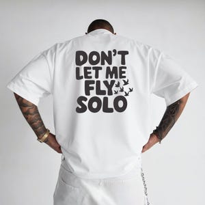 Puede incluir: Camiseta blanca con el texto "DON'T LET ME FLY SOLO" en letras negras. El diseño incluye una bandada de pájaros estilizados. La camiseta tiene mangas cortas y un corte holgado.