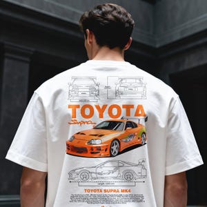 May include: White t-shirt featuring an orange Toyota Supra MK4 graphic. The design includes technical drawings, the Toyota logo, and the word "Supra." The car graphic is detailed with a spoiler and racing stripes.