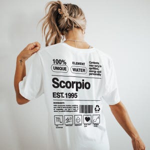 Puede incluir: Camiseta blanca con la palabra "Scorpio" en negrita negra, junto con el texto "EST. 1995". La camiseta también presenta las palabras "100% UNIQUE ELEMENT WATER" y una lista de ingredientes. La camiseta es una prenda única con temática zodiacal.