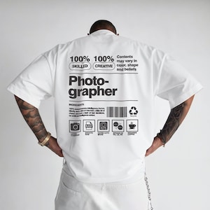 Może przedstawiać: Biała koszulka z napisem "Photographer" wydrukowanym dużymi czarnymi literami. Koszulka zawiera również tekst "100% Skilled, 100% Creative" oraz listę składników. Koszulka ma okrągły dekolt i krótkie rękawy.