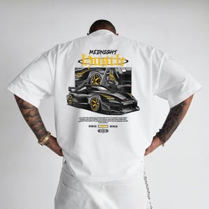 May include: White t-shirt with a graphic of a black sports car with gold accents. The design includes the text "MIDNIGHT Hustle" in a stylized font. Additional text is present below the car graphic.
