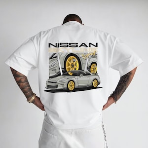 May include: White short-sleeve t-shirt featuring a graphic of a silver Nissan Skyline car with gold wheels. The text "NISSAN SKYLINE Legend" is printed above the car graphic in black and gold. The shirt is a casual style.