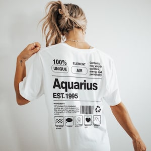 May include: White t-shirt with the word "Aquarius" in large black letters. The shirt also features the text "EST. 1995" and other text about the Aquarius zodiac sign. The shirt is a unique and stylish piece of apparel.