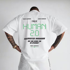 以下が含まれることがあります： 緑色の「EVOLVE HUMAN 2.0」の文字が入った白い半袖シャツ。「AUGMENTED HUMANOID」と「ARE YOU READY FOR AN UPGRADE?」のフレーズも黒で表示されています。「COGNITIVE NEUROSCIENCE」の文字がメインテキストの上に印刷されています。