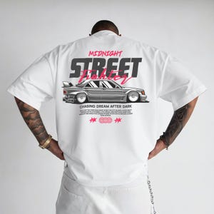 Puede incluir: Camiseta blanca de gran tamaño con un diseño gráfico en la espalda. El diseño presenta las palabras "MIDNIGHT STREET FIGHTER" sobre una ilustración de un coche plateado. Debajo del coche está el texto "CHASING DREAM AFTER DARK".