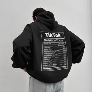 May include: Black hoodie with a white graphic on the back that reads "TikTok Nutrition Facts." The graphic lists values for creativity, screen time, social media engagement, and other categories. The hoodie has a hood and long sleeves.