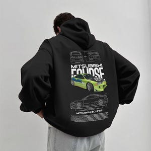 May include: Black hooded sweatshirt featuring a graphic of a Mitsubishi Eclipse car in green and yellow. The design includes car schematics and the text "MITSUBISHI ECLIPSE". The sweatshirt has a relaxed fit and a drawstring hood.