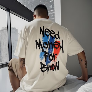 May include: A white t-shirt with the text "Need Money For BMW" in black, with blue and red brushstroke accents. The shirt is a casual style.