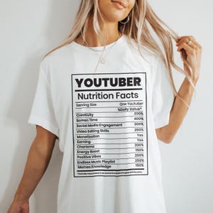 Puede incluir: Camiseta blanca con un gráfico negro que dice "YOUTUBER Nutrition Facts". El gráfico enumera atributos como creatividad y tiempo de pantalla, con valores porcentuales. La camiseta es de manga corta.