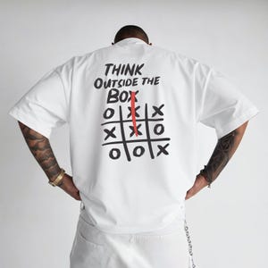 Może przedstawiać: Biała koszulka z napisem "THINK OUTSIDE THE BOX" nad siatką do gry w kółko i krzyżyk. Siatka ma czerwoną linię pionową. Koszulka ma krótkie rękawy.