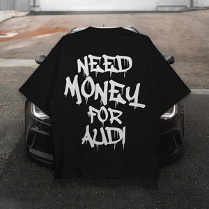 May include: Black oversized t-shirt with the text "NEED MONEY FOR AUDI" in white, dripping font. The shirt is displayed over the hood of a black car, showcasing the back of the shirt.